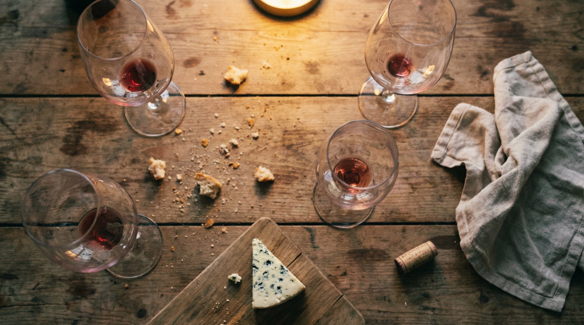 Top-down view of wine glasses and a wedge of blue cheese on a wooden table, bread crumbs and a cork scattered around